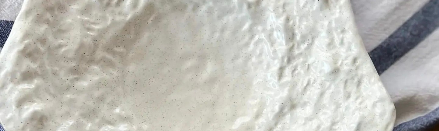 Ceramic plate with an organic shape and a white textured surface on a wooden table, next to a fabric napkin and a branch with cotton flowers.